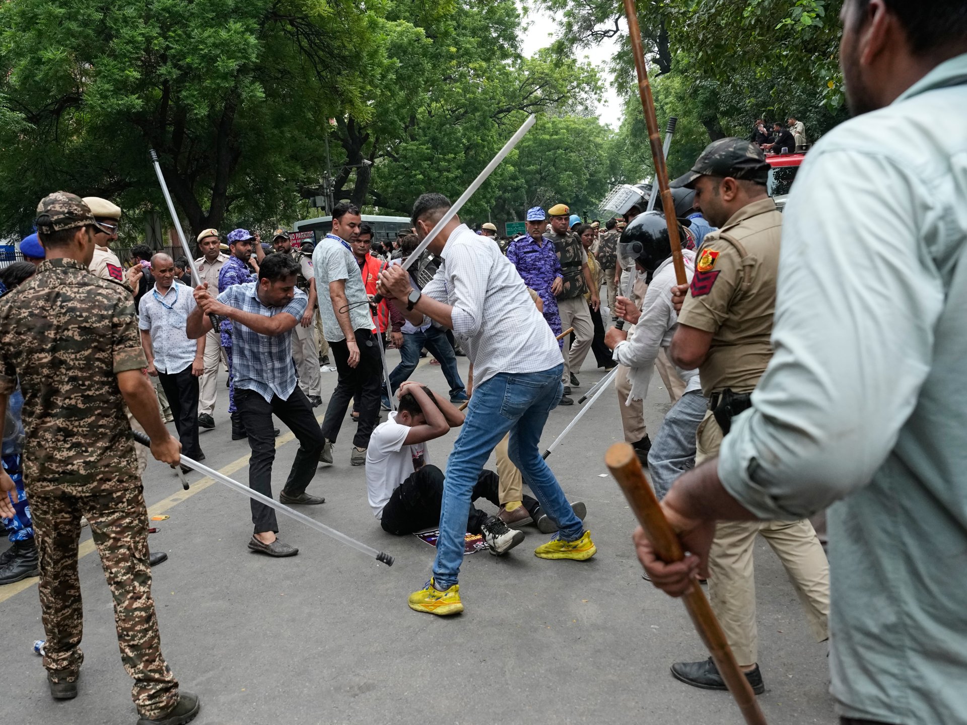 Police and paramilitary personnel with lathis surrounding a seated protester who is covering his head